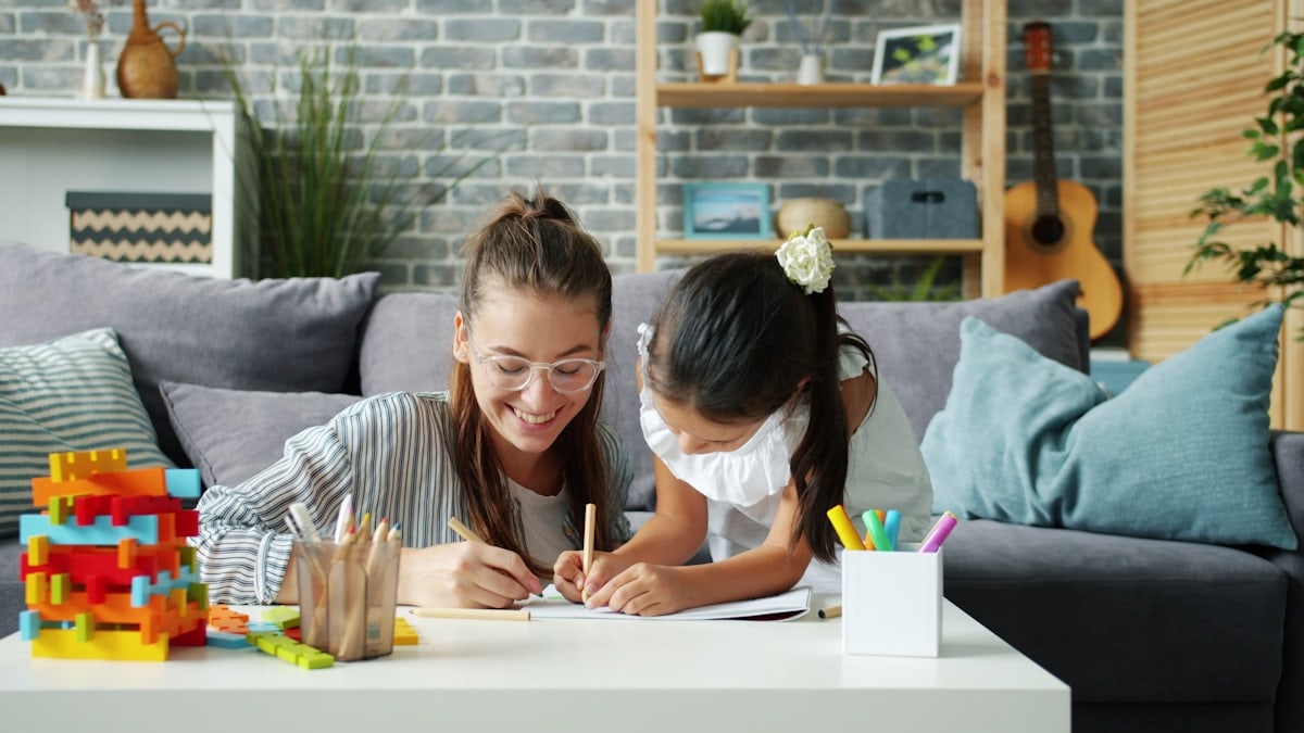 Happy little girl is drawing at home with caring mother using pencils having fun in modern apartment. Creative leisure activity and childhood concept.
